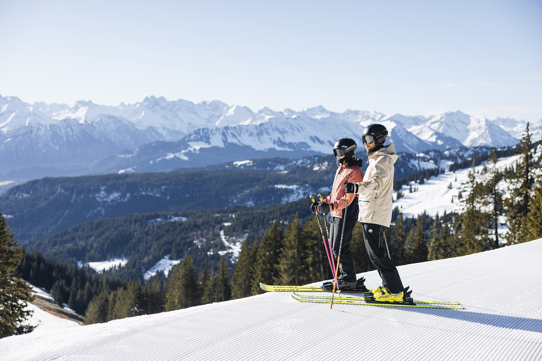 Zwei Skifahrer stehen vor einem Abhang mit Bergausblick.