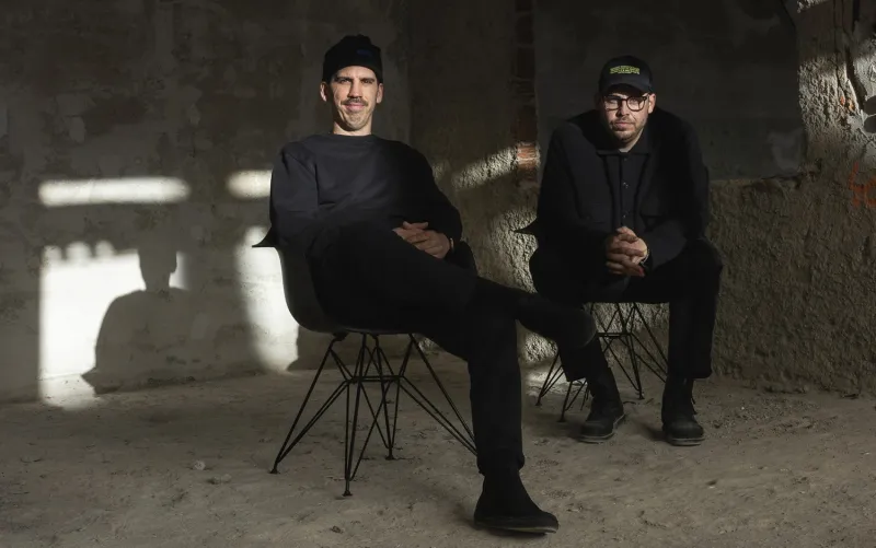 Die Gründer Tom (links) und Philipp Herzhoff in einem Rohbau-Raum ganz in schwarz gekleidet und sitzend.