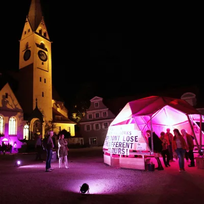 Der Sankt-Mang Platz bei Nacht, mit beleuchteter Kirche und einem pink beleuchteten KunstNacht Zelt im Vordergrund